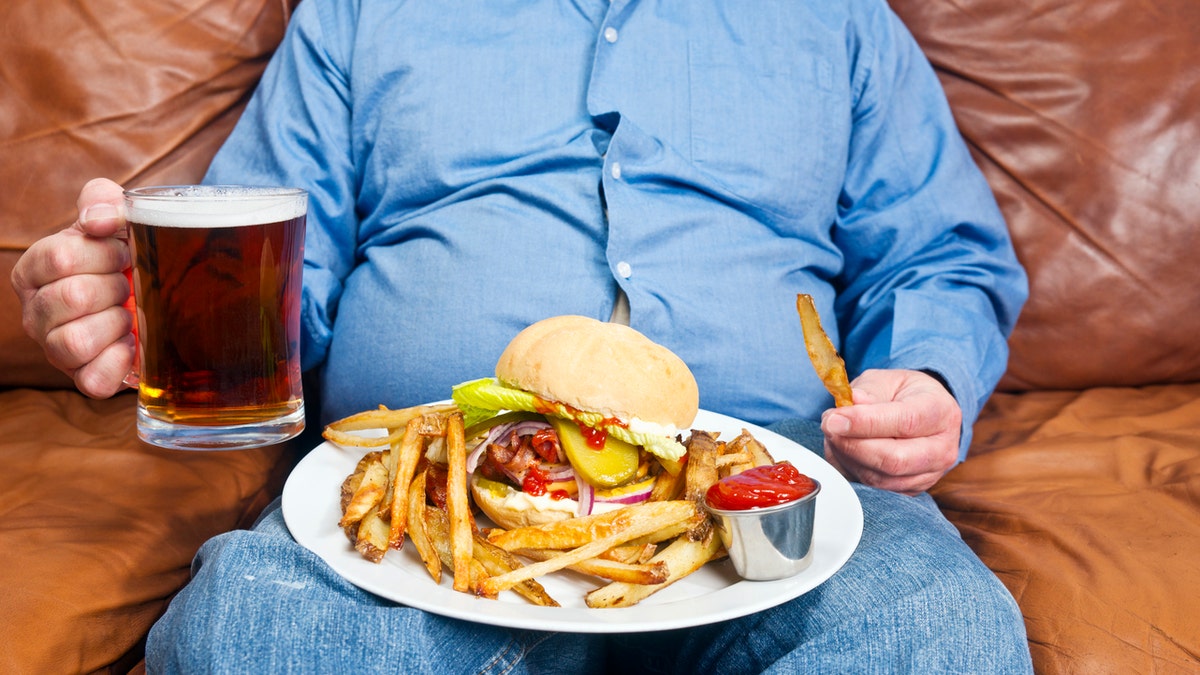 A photo of an overweight man sitting on an old couch with a very large unhealthy meal on his lap and a pint of beer in his hand. Obesity is a major cause of diabetes.