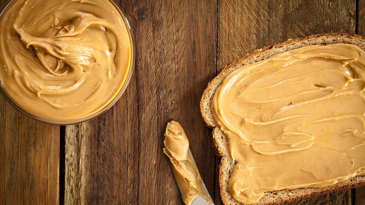 A bread slice sits on the right side of the image over a rustic wooden table with a table knife with peanut butter on top and a bowl full of peanut butter on left top, shot was made with a Canon EOS 5D Mark II directly from above and peanut butter is spread on bread top.