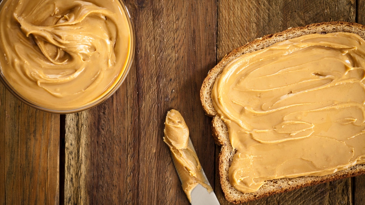 A bread slice sits on the right side of the image over a rustic wooden table with a table knife with peanut butter on top and a bowl full of peanut butter on left top, shot was made with a Canon EOS 5D Mark II directly from above and peanut butter is spread on bread top.