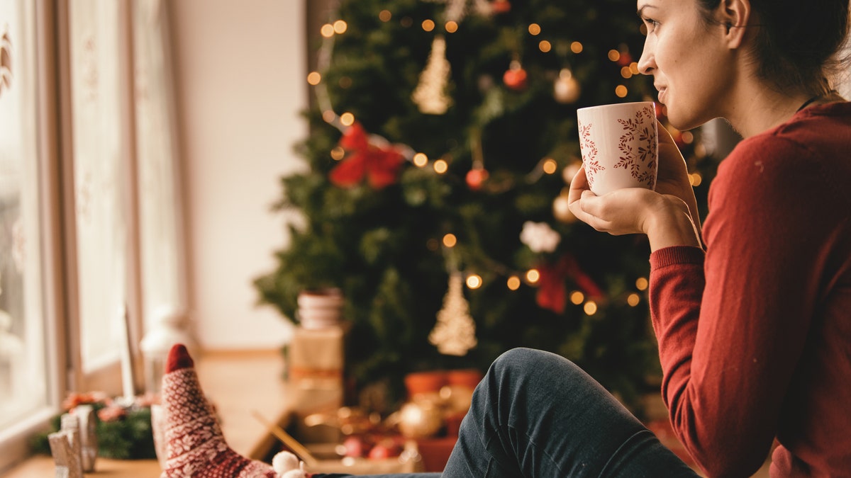 Young woman drinking tea by the Christmas tree, looking through window.