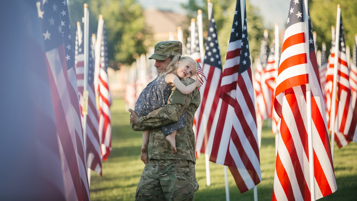 3 year old girl running with her military dad who is wearing an American Army uniform in a field of American flags.