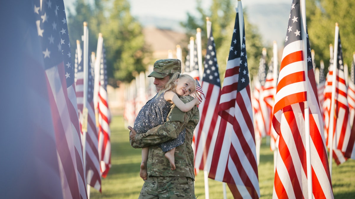 3 year old girl running with her military dad who is wearing an American Army uniform in a field of American flags.