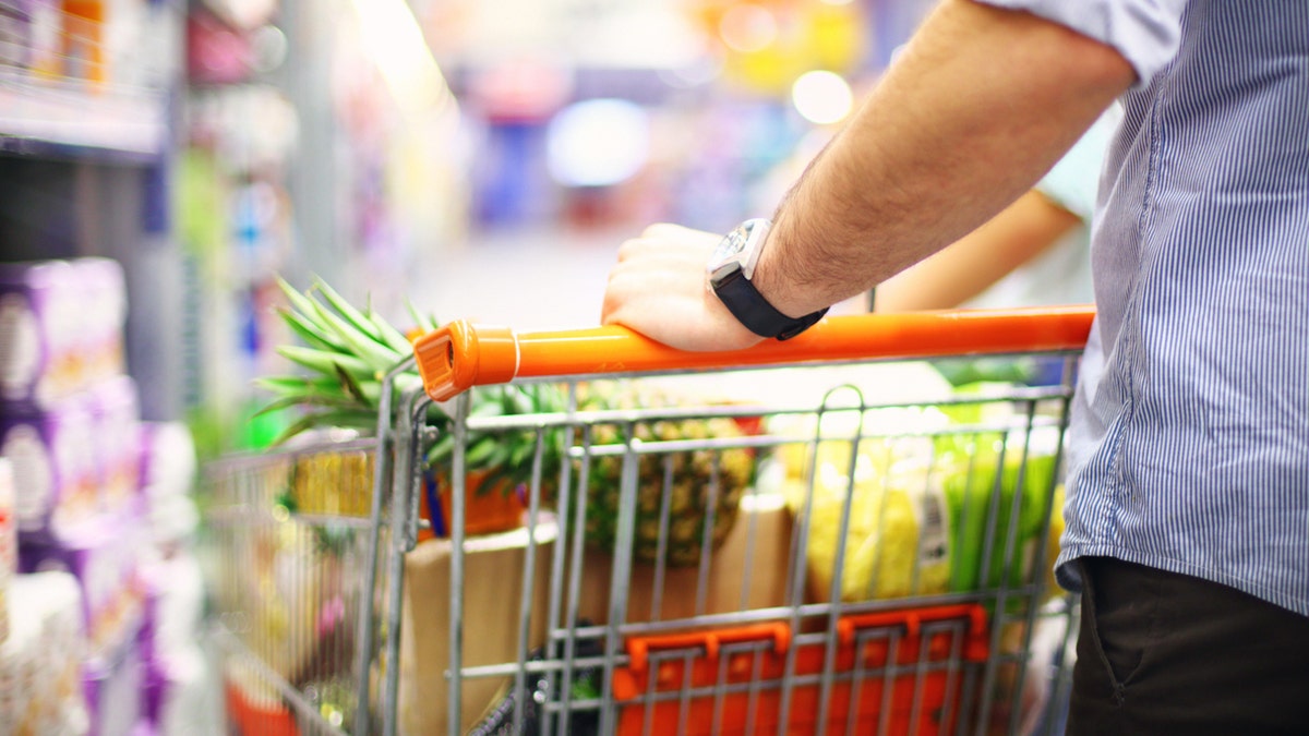 Rear view of unrecognizable mid-aged man in supermarket.He's pushing shopping car almost fully packed with groceries.Shelves with products stretch deep in background out of focus. He's wearing light blue striped shirt with rolled back sleeves,brown pants and a wristwatch.