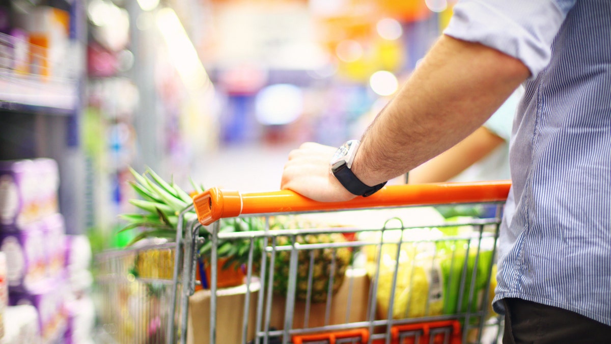 Rear view of unrecognizable mid-aged man in supermarket.He's pushing shopping car almost fully packed with groceries.Shelves with products stretch deep in background out of focus. He's wearing light blue striped shirt with rolled back sleeves,brown pants and a wristwatch.