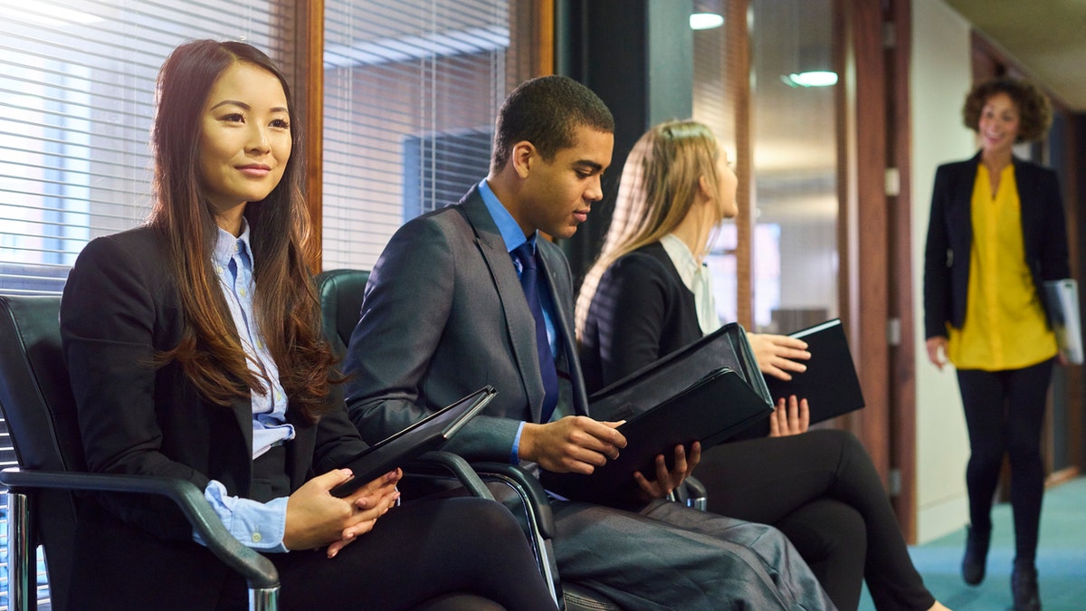 three interview candidates wait nervously in the reception of an office building. a member of the interview panel can be seen walking towards them.