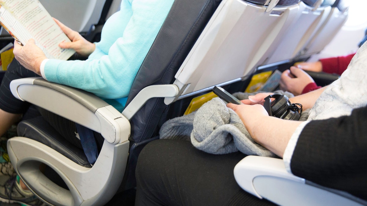 In this real situation a row of unidentifiable passengers are sitting in their seats on an airplane. The seating is tight. Their legs are touching the seats in front of them.