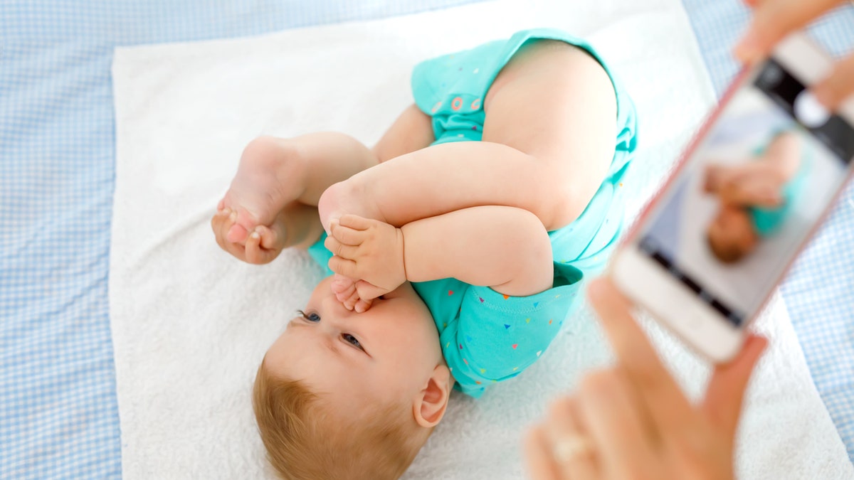 Parent taking photo of a baby with smartphone. Adorable newborn child taking foot in mouth. sucking feet. Digital family memories