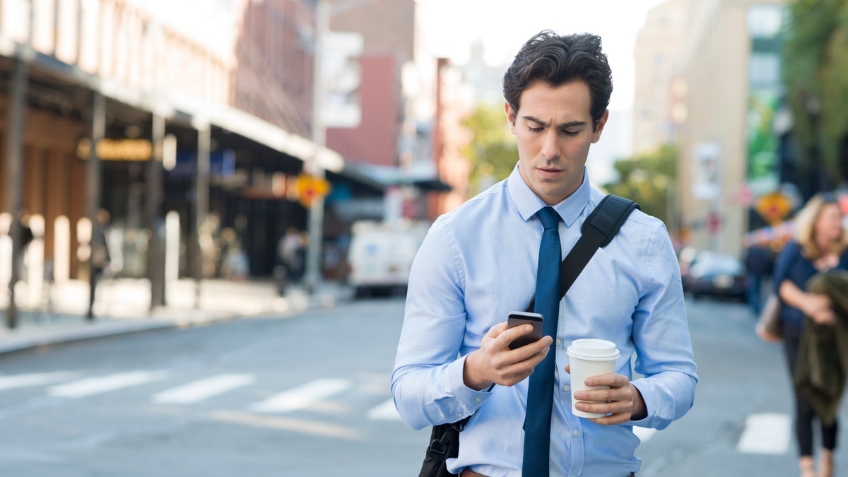 Businessman using smartphone and holding paper cup ina urban scene. Worried businessman in walking on the road and messaging with phone. Young man text messaging through cell phone while walking on the road in the city centre.