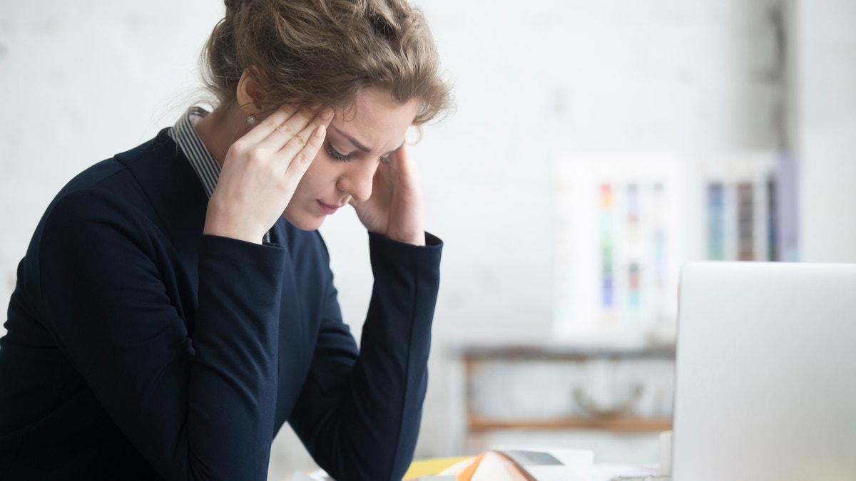 Portrait of young stressed woman sitting at home office desk in front of laptop, touching head with frustrated facial expression, having headache, overworked or depressed