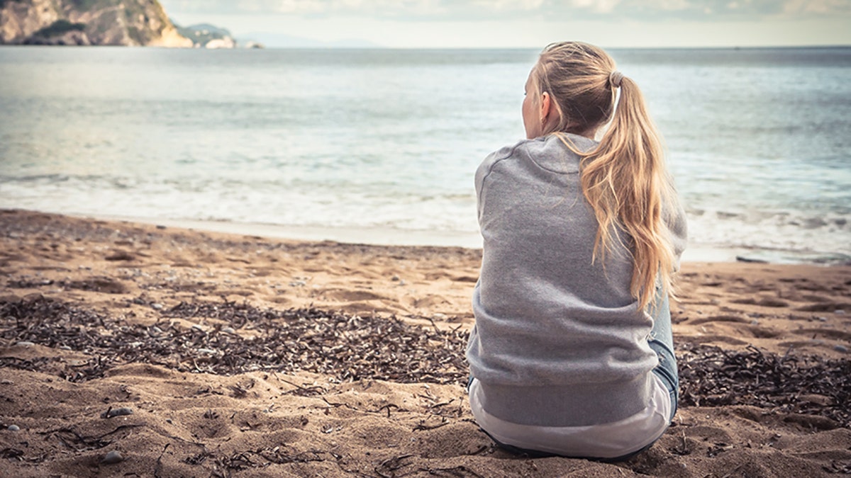Pensive lonely young woman tourist sitting on beach hugging her knees and looking into the distance with hope