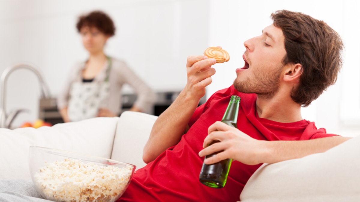 A man lying with snacks and beer on a couch and his busy wife in kitchen