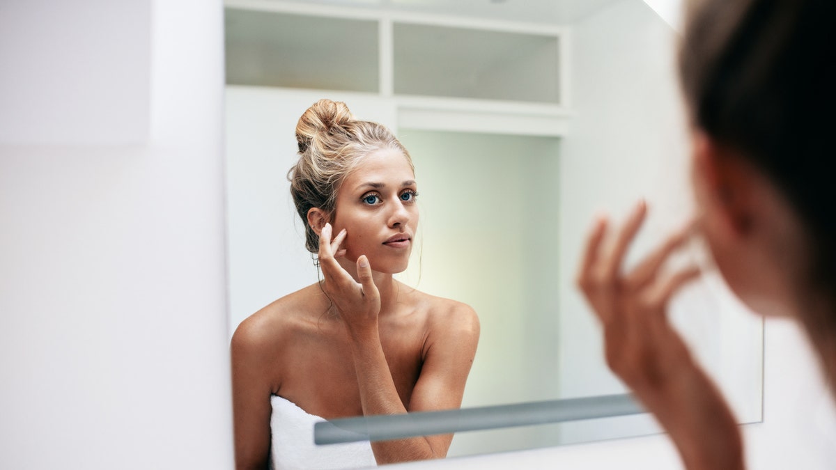 Beautiful young woman standing in the bathroom. Female looking into the mirror and touching her face skin.