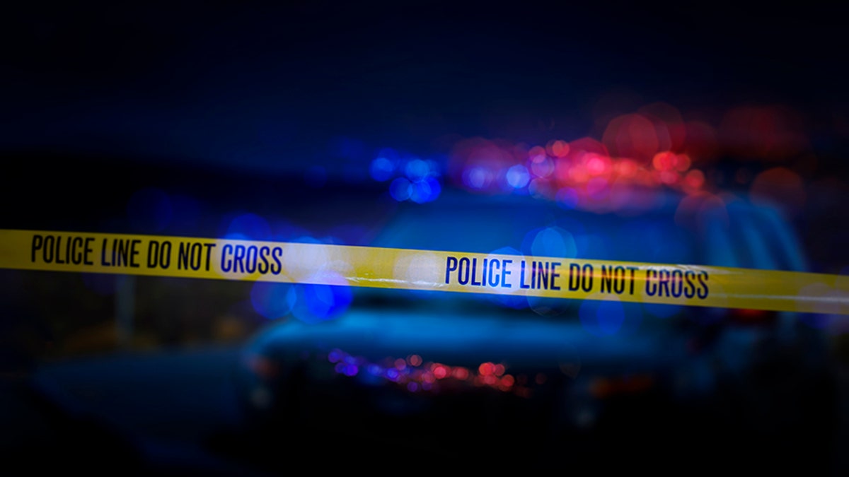 A stock photo of a Police Line "Do Not Cross" caution tape with a defocused police car with sirens flashing red and blue.