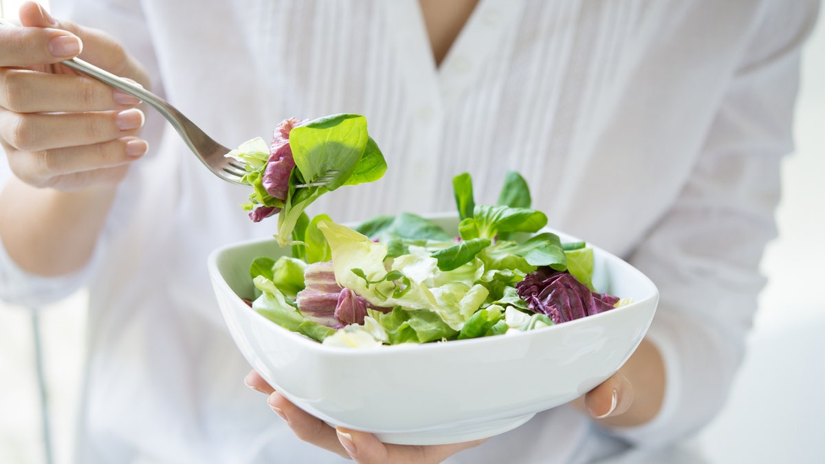 Close-up shot of a woman holding a plate of fresh green salad in the beautiful morning light.  She is holding a fork and about to eat the vegetarian food.  Healthy eating and diet concept.  Shallow depth of field with focus on the fork.