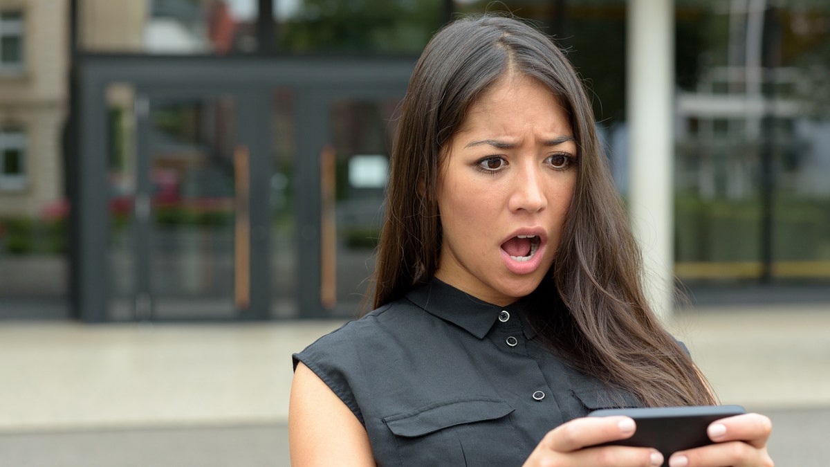 Young woman reacting in horror to an sms, text message or email on her mobile phone as she stands outdoors in a quiet urban street, close up head and shoulders