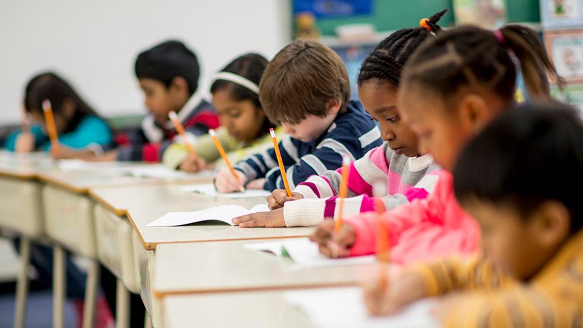 A multi-ethnic group of elementary school children are indoors in a classroom. They are wearing casual clothing. The students are sitting at their desks and writing with pencils.