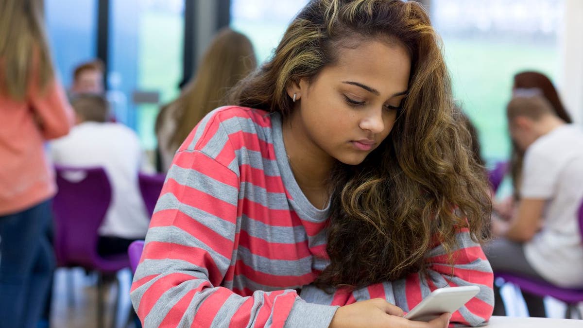 Female student sitting on her own at school. She has a smartphone in her hand and a stressed expression on her face.