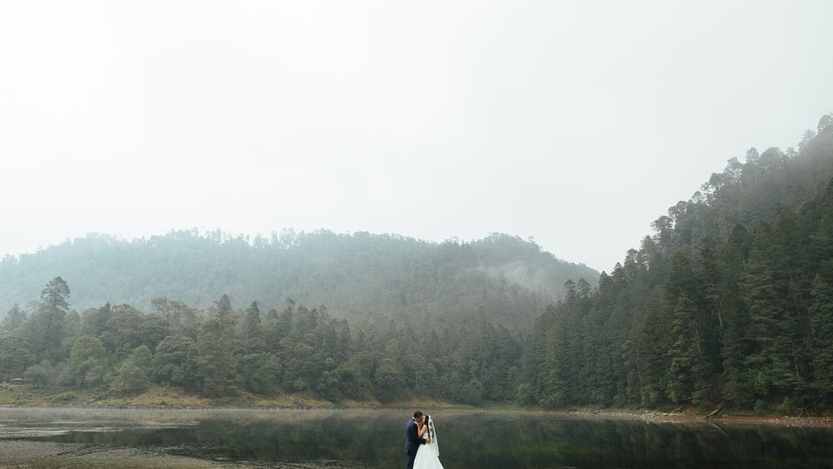 A romantic latin newlywed couple standing and kissing outdoors in a foggy day in a horizontal full length shot.