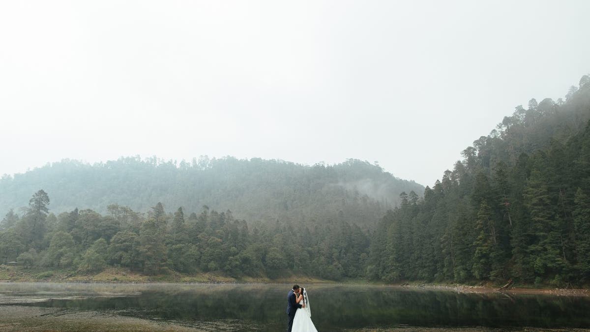 A romantic latin newlywed couple standing and kissing outdoors in a foggy day in a horizontal full length shot.