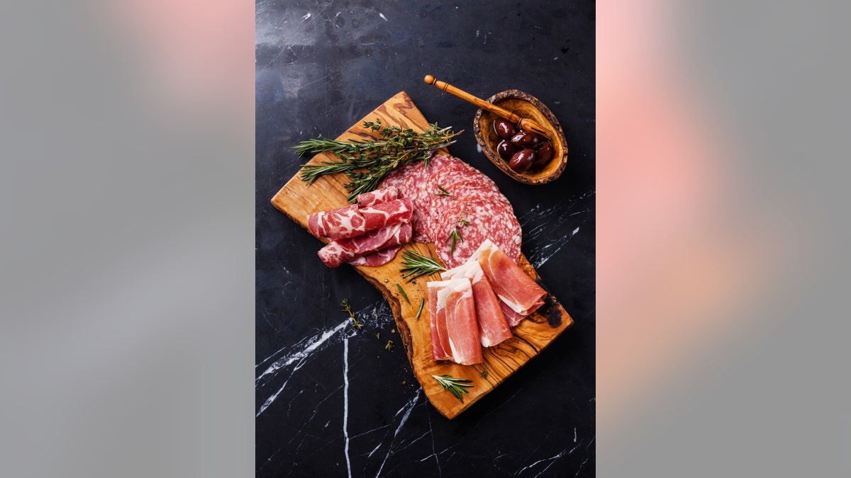 Sliced prosciutto di Parma on wooden board with salami and rosemary on black marble background