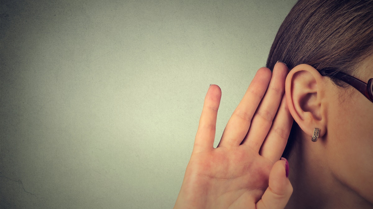 Woman holds her hand near ear and listens carefully isolated on gray wall background