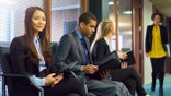 three interview candidates wait nervously in the reception of an office building. a member of the interview panel can be seen walking towards them.