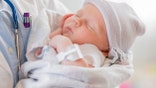 A Caucasian female infant is asleep in the arms of a pediatrician. The baby is swaddled in a blanket and is wearing a warm cap.