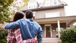 A couple hugging one another in front of a big frame house.