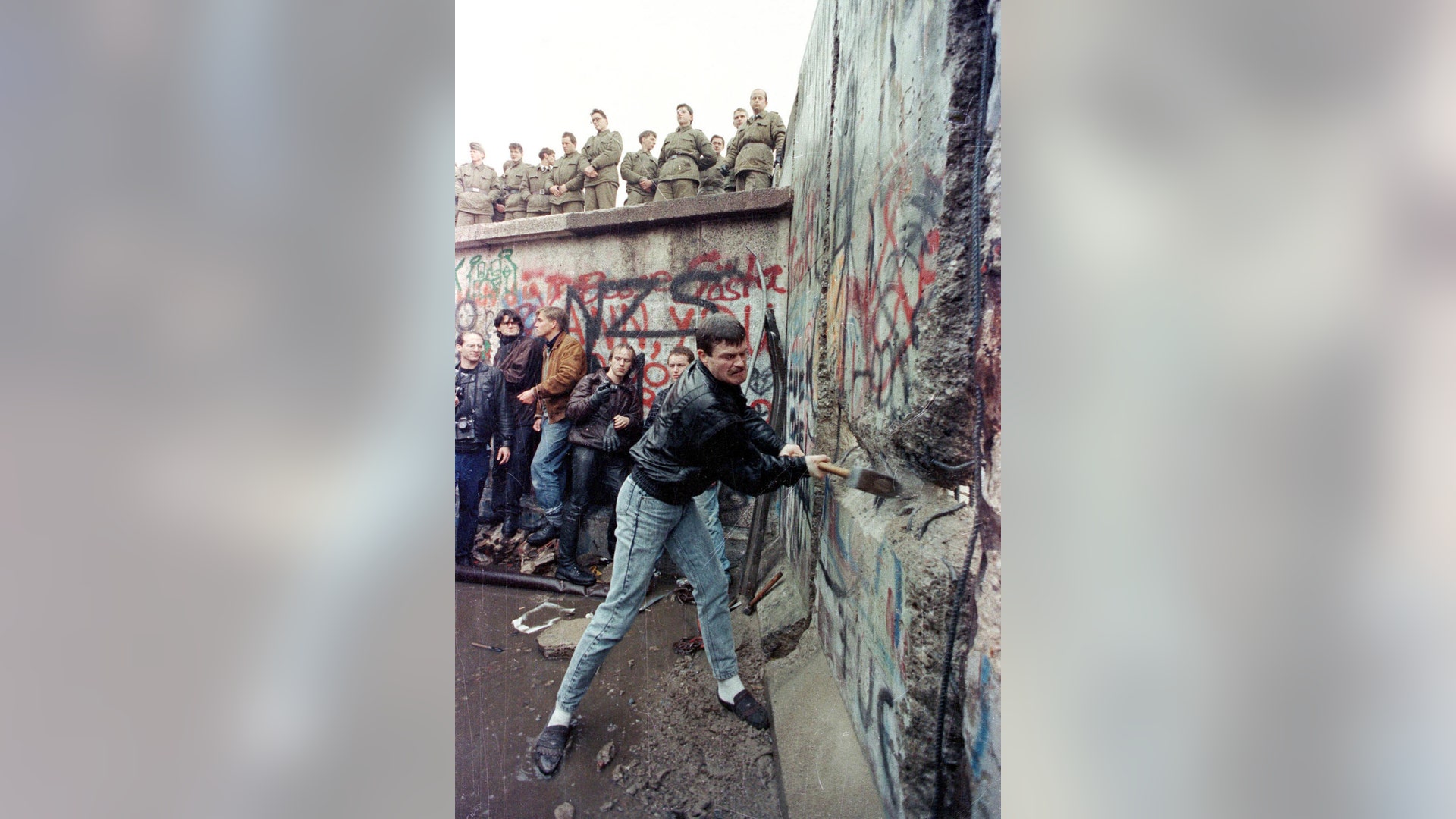 A demonstrator hits the Berlin Wall as East Berlin border guards look on from above the Brandenburg Gate, November 11, 1989