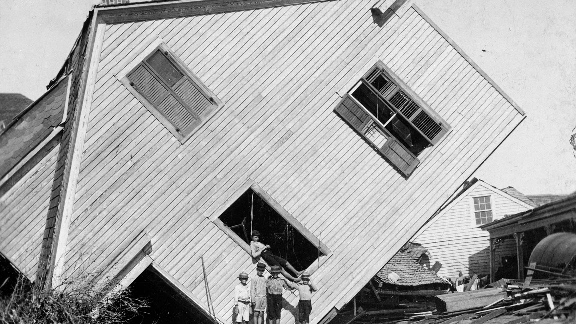 A house tipped on side with several boys standing in front after a flood in Galveston, Texas, 1900