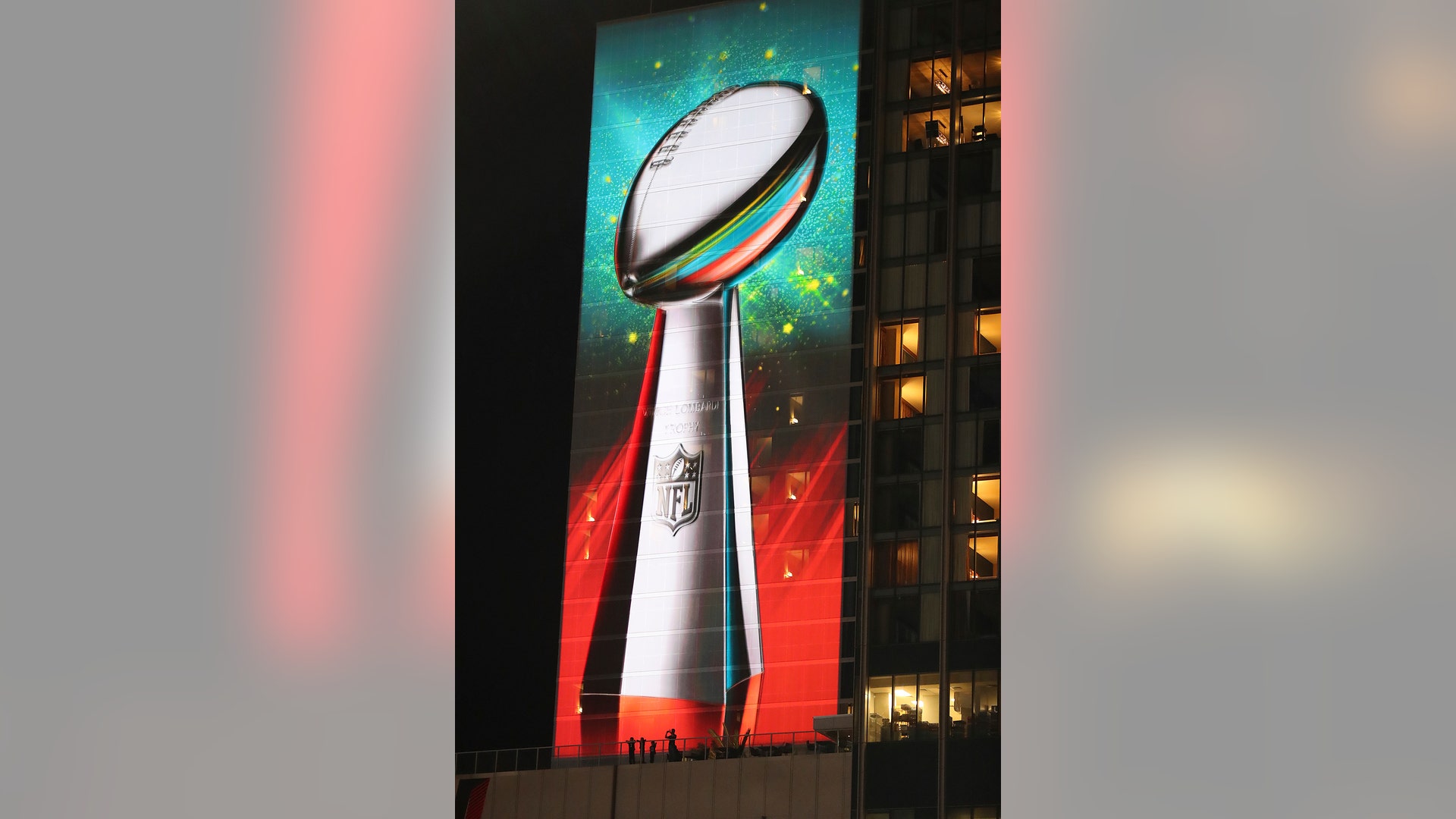 Football fans are dwarfed by a giant projection of the Lombardi trophy on the side of a hotel during Super Bowl LIVE. 