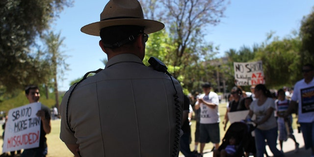 PHOENIX - MAY 01: A police officer watches demonstrators protest Arizona's new immigration enforcement law during a May Day rally at the state capitol building on May 1, 2010 in Phoenix, Arizona. Thousands of people in cities across the United States protested the immigration law, which critics have said could encourage racial profiling against Hispanics by police officers. (Photo by John Moore/Getty Images)