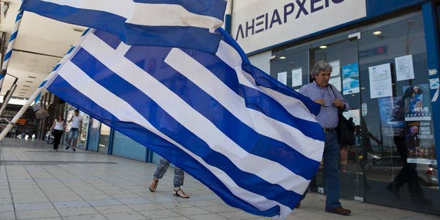 Pedestrians walk in front of a civil registry office as Greek flags are displayed for sale in the foreground at Athens' main port of Piraeus on Monday, June 6, 2016. Bailout lenders on Monday were reviewing new Greek austerity measures demanded to release the next installments of rescue loans for Athens. (AP Photo/Petros Giannakouris)