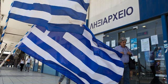 Pedestrians walk in front of a civil registry office as Greek flags are displayed for sale in the foreground at Athens' main port of Piraeus on Monday, June 6, 2016. Bailout lenders on Monday were reviewing new Greek austerity measures demanded to release the next installments of rescue loans for Athens. (AP Photo/Petros Giannakouris)