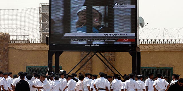 Aug. 15: Egyptian police officers watch 83-year-old former Egyptian president Hosni Mubarak on a bed in the defendants' cage from the courtroom live on a large TV screen outside the Police Military Academy complex in Cairo, Egypt.