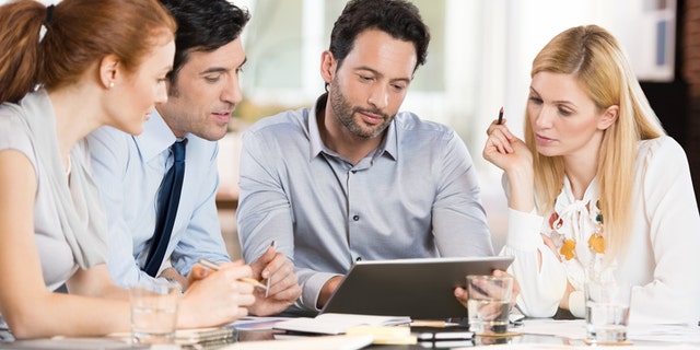 Confident businessmen and businesswomen sitting and working on tablet during a meeting. Business people sitting around the desk in a meeting and watching digital tablet. Portrait of happy colleagues looking at laptop screen in office.