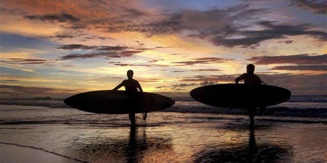 Two surfers carrying their boards are silhouetted against the sunset at Kuta Beach in Bali, Indonesia.