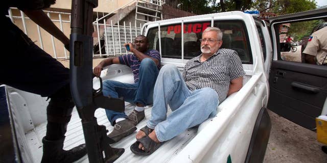 FILE - In this Friday, Sept. 5, 2014, file photo, Michael Geilenfeld waits in handcuffs as the manager of his orphanage sits with him in the back of a police truck outside the St. Joseph's Home For Boys after police closed it down in the Delmas area of Port-au-Prince, Haiti. Hearts With Haiti, a nonprofit that raises money for the orphanage, and Geilenfeld have accused activist Paul Kendrick of spreading falsehoods that have cost the charity more than $2 million in donations. A jury that was seated Monday, July 6, 2015 for the trail. (AP Photo/Dieu Nalio Chery, File)