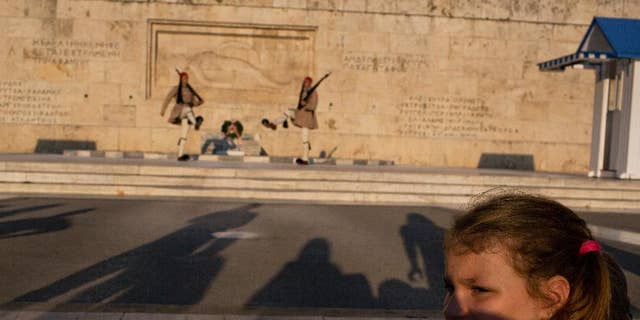 A visitor looks away from the changing of the guard ceremony at Greece's parliament to watch a small number of protesters across the street, in central Athens on Tuesday, July 5, 2016. The tiny anti-government rally was held to mark a year since Greeks voted against bailout measures in a referendum. Shortly after the vote, Greece reaching a third bailout deal with international rescue creditors. (AP Photo/Petros Giannakouris)