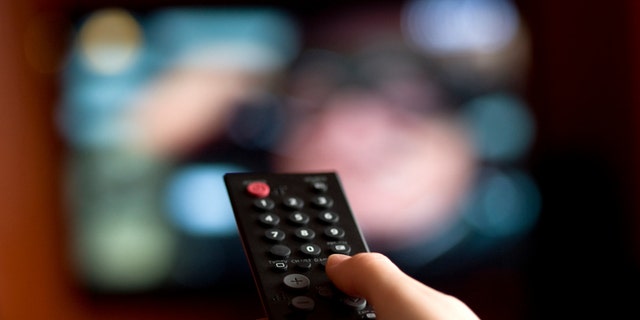 Focused on the hand holding a remote control with a television in the background.