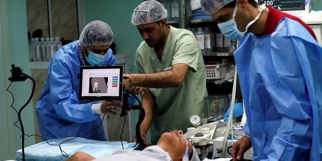 A patient lies on a bed as a technician adjusts a screen before the start of a Proximie surgery, with doctor Ghassan Abu Sitta in Beirut guiding Palestinian surgeon Hafez Abu Khousa in the operating room at Al Awda Hospital in the northern Gaza Strip