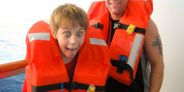 Father and his son put on life vests during cruise ship safety drill before departing. Ocean in background. Son makes silly face just as mom snaps the vacation photo.