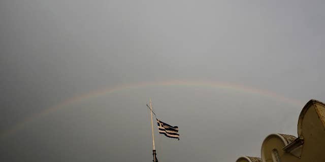 A Greek flag flies on top of the municipality market with rainbow on the background on Kos island, Greece, Friday, May 29, 2015. Bank deposits in Greece have hit their lowest in more than a decade, according to data released Friday, as Greeks concerned about the painfully slow pace of their government's talks with bailout creditors withdrew savings in an orderly but steady flow.(AP Photo/Petros Giannakouris)