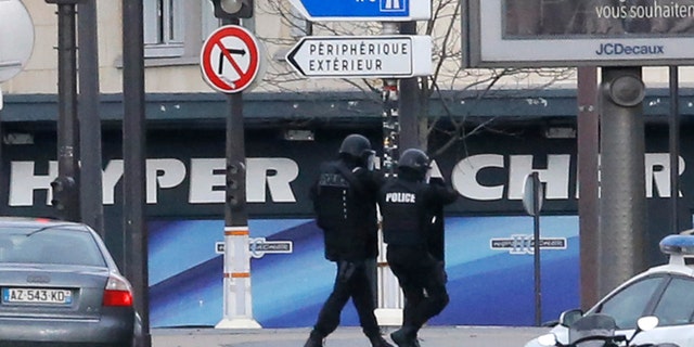 Jan.9, 2015: Police officers walk in front of a hostage-taking situation at a kosher market, seen in blue in background, in Paris, Friday . A police official says the man who has taken at least five people hostage in a kosher market in Paris appears linked to the newsroom massacre earlier this week that left 12 people dead.