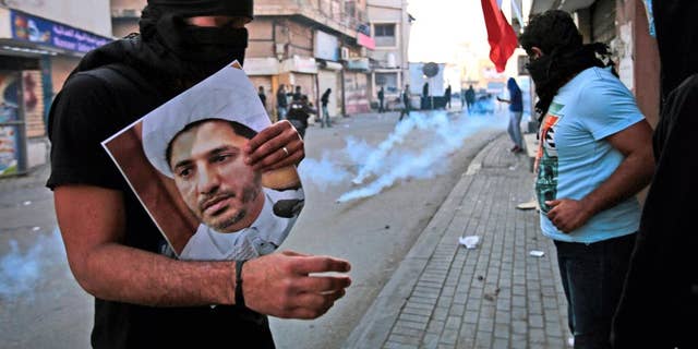 FILE- In this Jan. 3, 2015 file photo, a masked Bahraini anti-government protester holds a picture of jailed Shiite cleric Sheik Ali Salman, the head of the opposition al-Wefaq political association, as riot police fire tear gas canisters during clashes in Bilad Al Qadeem, Bahrain. On Tuesday, June, 14, 2016, Bahrain said it has suspended all activities by Al-Wefaq, the largest Shiite opposition political group, and frozen its assets amid a widening crackdown on dissent, five years after the country’s Arab Spring protests. A statement from the Justice and Islamic Affairs Ministry carried on the state-run Bahrain News Agency said a court made the decision to suspend Al-Wefaq to “safeguard the security of the kingdom.” (AP Photo/Hasan Jamali, File)