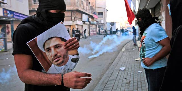FILE- In this Jan. 3, 2015 file photo, a masked Bahraini anti-government protester holds a picture of jailed Shiite cleric Sheik Ali Salman, the head of the opposition al-Wefaq political association, as riot police fire tear gas canisters during clashes in Bilad Al Qadeem, Bahrain. On Tuesday, June, 14, 2016, Bahrain said it has suspended all activities by Al-Wefaq, the largest Shiite opposition political group, and frozen its assets amid a widening crackdown on dissent, five years after the country’s Arab Spring protests. A statement from the Justice and Islamic Affairs Ministry carried on the state-run Bahrain News Agency said a court made the decision to suspend Al-Wefaq to “safeguard the security of the kingdom.” (AP Photo/Hasan Jamali, File)