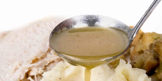 A spoonful of gravy being poured over mashed potatoes. Turkey and dressing in the background.