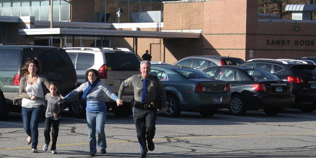 Dec. 14, 2012: In this file photo provided by the Newtown Bee, a police officer leads two women and a child from Sandy Hook Elementary School.