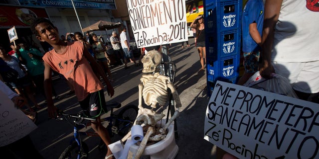 A plastic skeleton sits on a toilet with a banner attached that reads in Portuguese; "I'm still waiting for sanitation, signed Ze Rocinha: 2014," alongside a doll that sits on a potty with a banner that reads in Portuguese: "They promised me sanitation, signed Ze Rocinha: 1960," during a demonstration organized by the pro-sanitation group Meu Rio at the Rocinha slum, in Rio de Janeiro, Brazil, Saturday, Feb. 22, 2014. Ze Rocinha is a character invented by Rocinha slum residents. Meu Rio aims to draw attention to the Olympic city’s massive sewage problem. Only 30 percent of Rio’s sewage is treated, and the rest flows raw into its waterways. (AP Photo/Silvia Izquierdo)