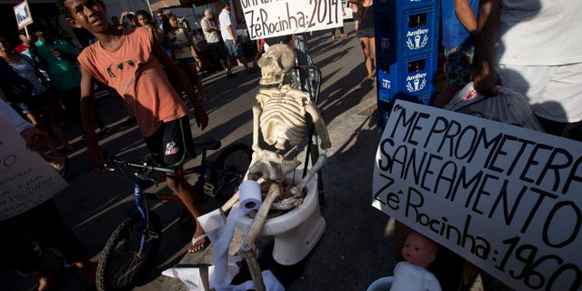 A plastic skeleton sits on a toilet with a banner attached that reads in Portuguese; "I'm still waiting for sanitation, signed Ze Rocinha: 2014," alongside a doll that sits on a potty with a banner that reads in Portuguese: "They promised me sanitation, signed Ze Rocinha: 1960," during a demonstration organized by the pro-sanitation group Meu Rio at the Rocinha slum, in Rio de Janeiro, Brazil, Saturday, Feb. 22, 2014. Ze Rocinha is a character invented by Rocinha slum residents. Meu Rio aims to draw attention to the Olympic city’s massive sewage problem. Only 30 percent of Rio’s sewage is treated, and the rest flows raw into its waterways. (AP Photo/Silvia Izquierdo)